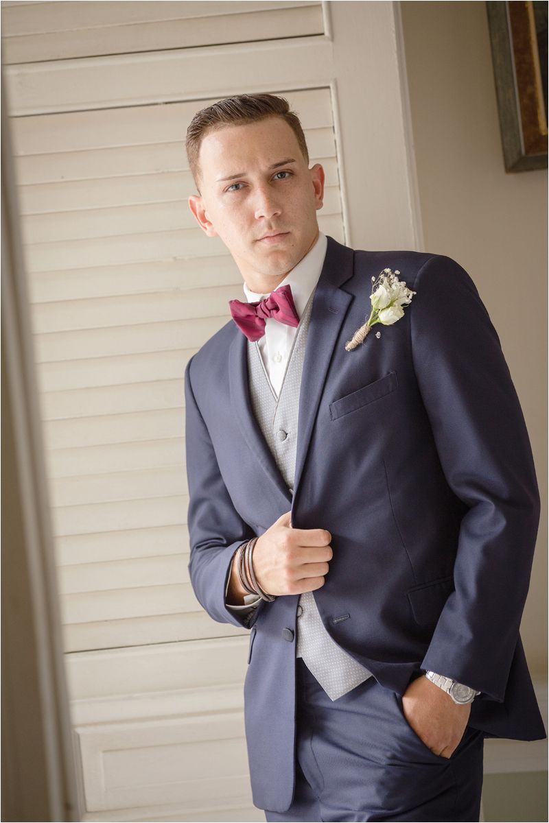 A groom in a navy suit with a burgundy bowtie and boutonniere poses in multiple formal portraits.