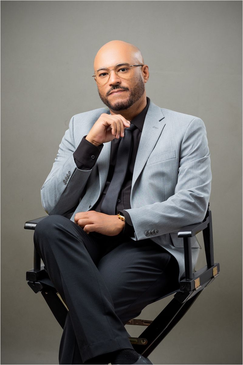 Professional studio portrait of someone seated in a director's chair wearing a light gray suit against neutral backdrop.
