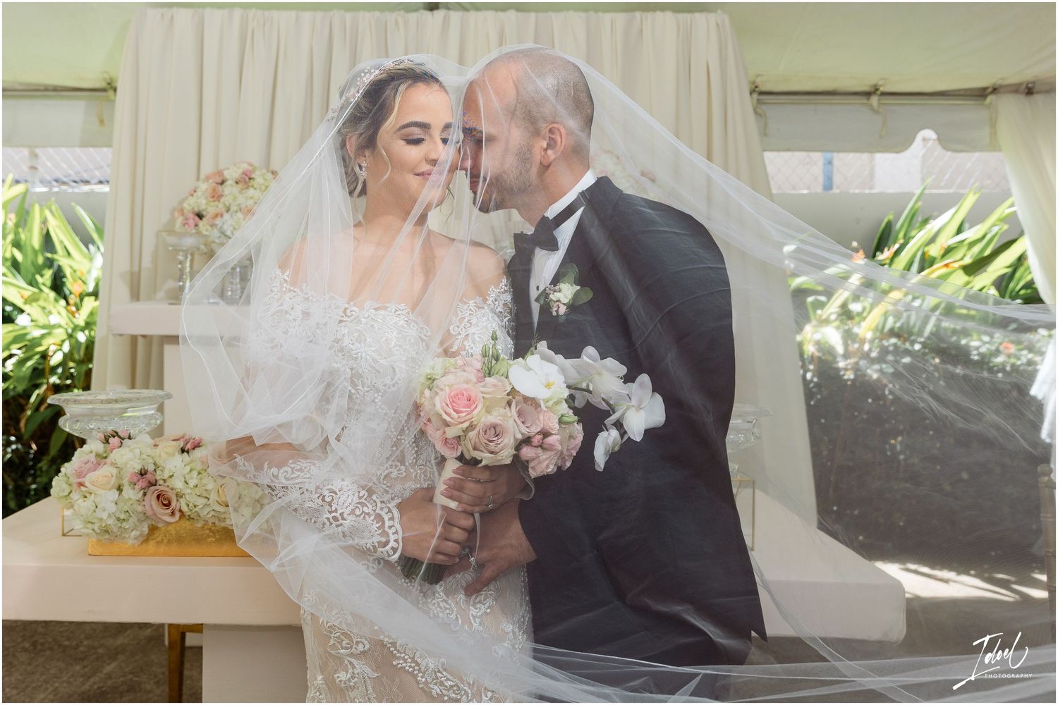 Romantic wedding moment captured under a cream-colored gazebo with pink floral bouquet and flowing veil.