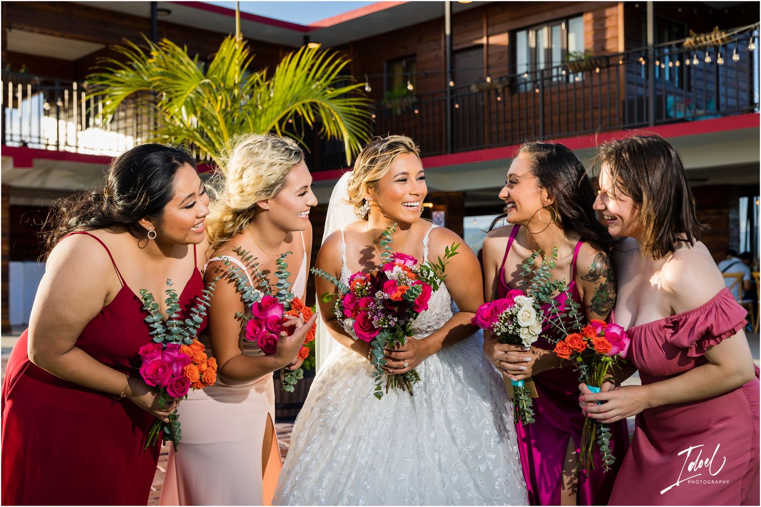 Candid moment of bridal party laughing together while holding pink bouquets against modern building.