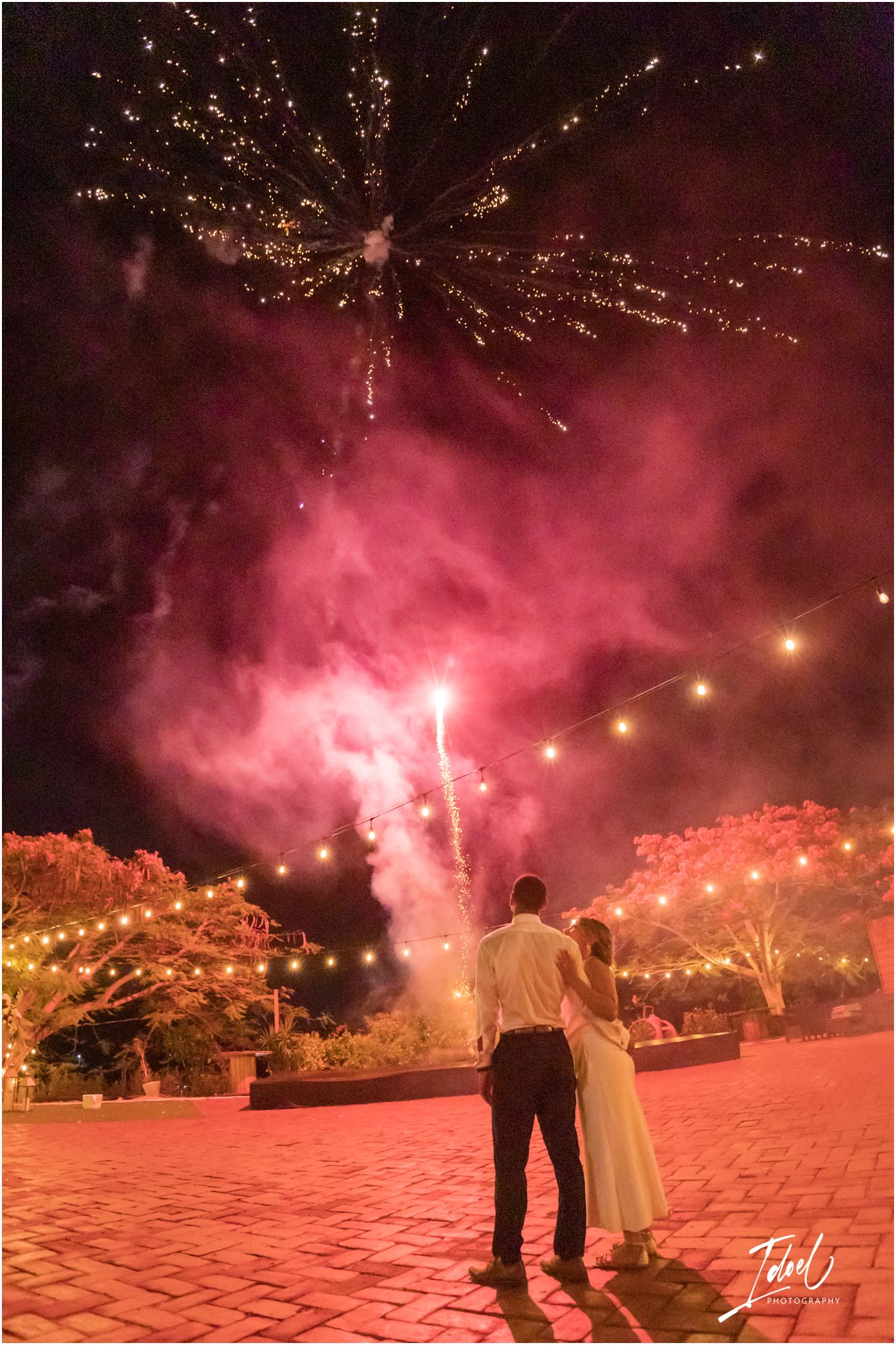 Wedding couple watching pink fireworks display against night sky with string lights below.