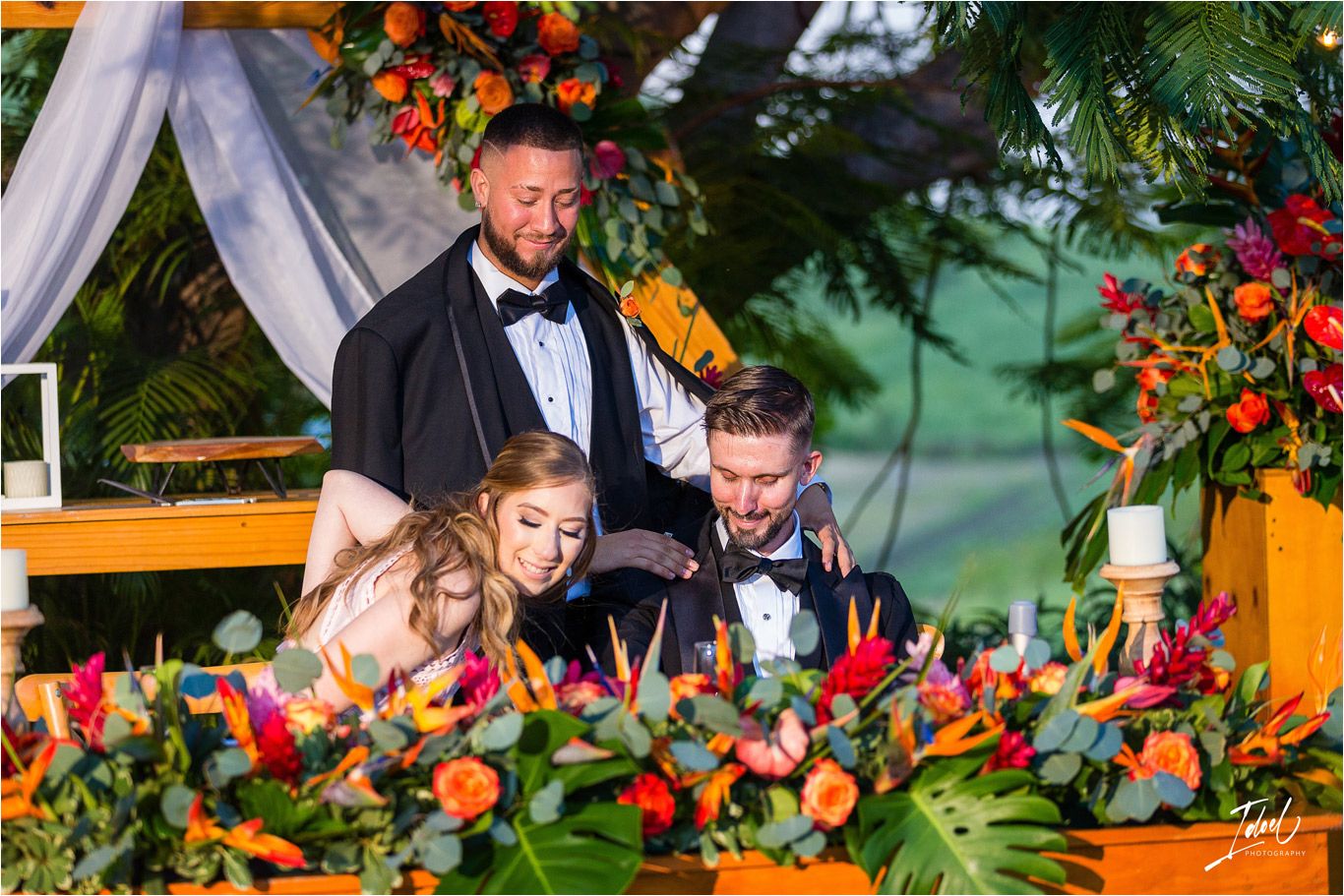 Wedding party shares laughter at reception table decorated with colorful flowers and greenery.
