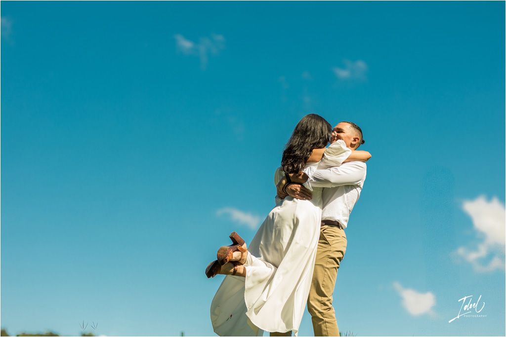 A couple share a loving embrace against a bright blue sky with white fluffy clouds on a sunny day.