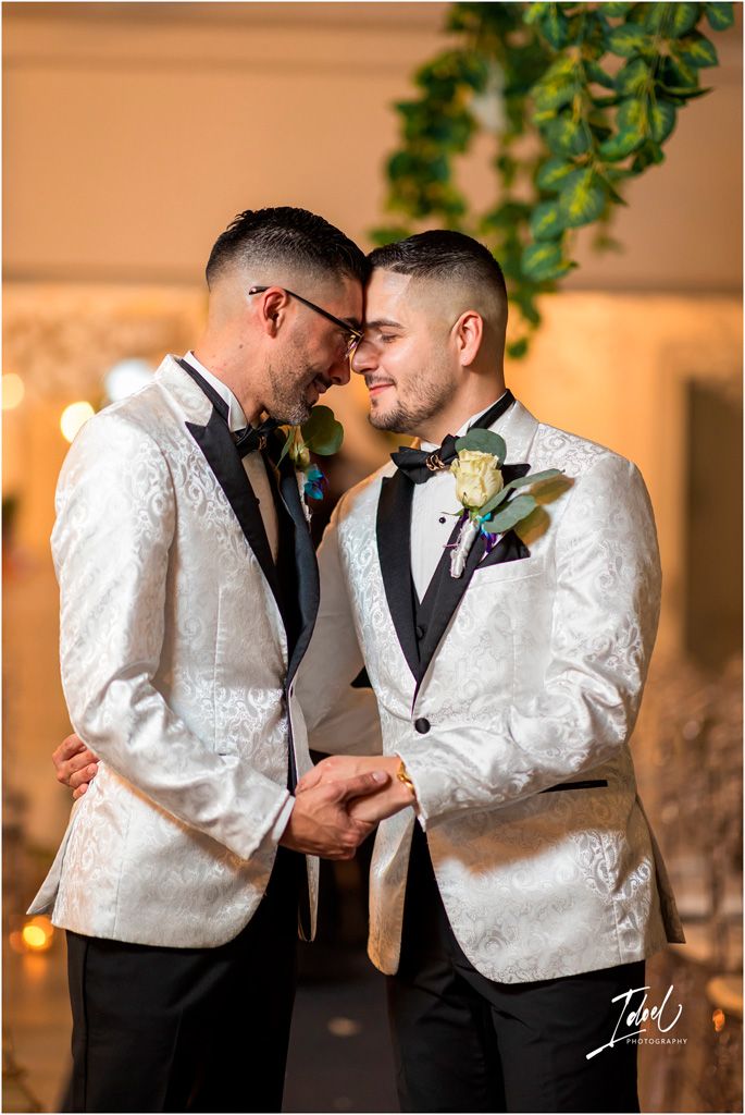 Two people in matching white tuxedo jackets and bow ties share an intimate moment at their wedding celebration.