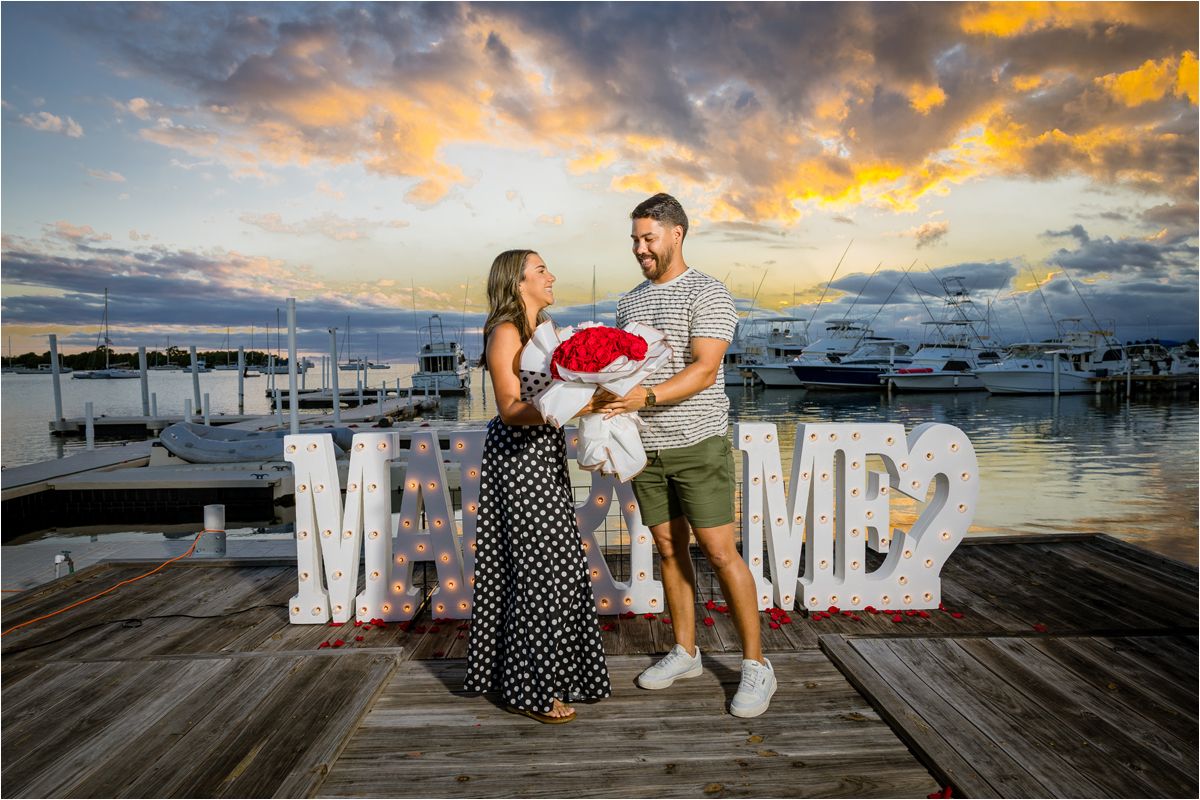 A couple stands on a marina dock at sunset with decorative 'MR' letters behind them as boats and clouds fill the background.