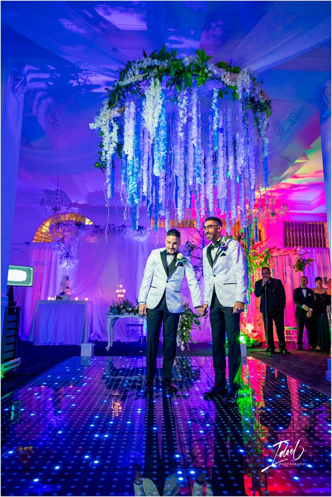 Two people in white tuxedos stand under an illuminated floral chandelier on a glowing dance floor with purple lighting.