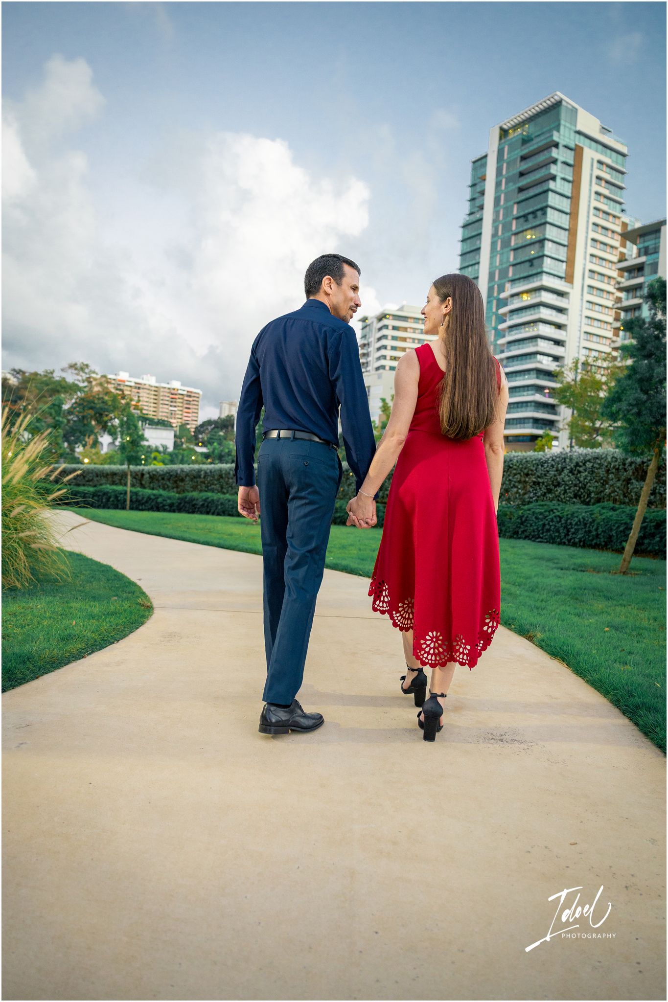 Couple holding hands walking on a curved path with city buildings and green hedges in the background.