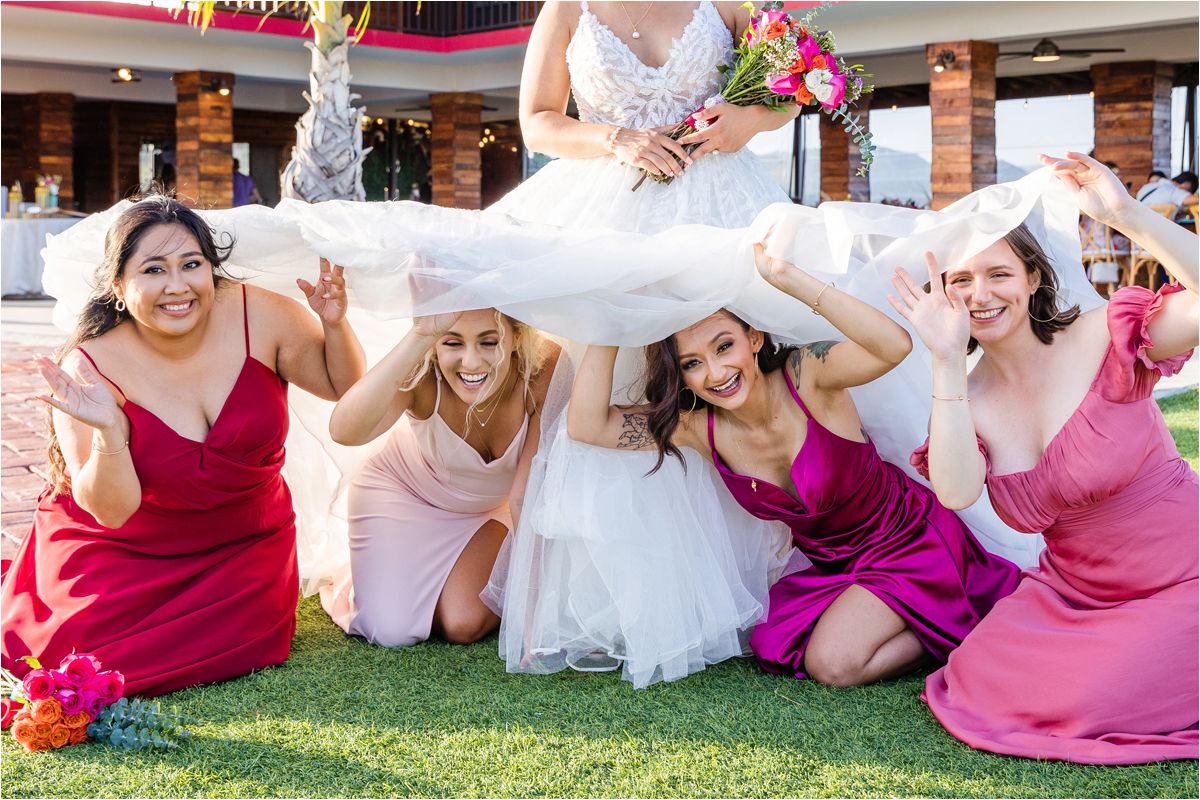 Wedding party in pink and burgundy dresses poses playfully on grass during outdoor photo session.