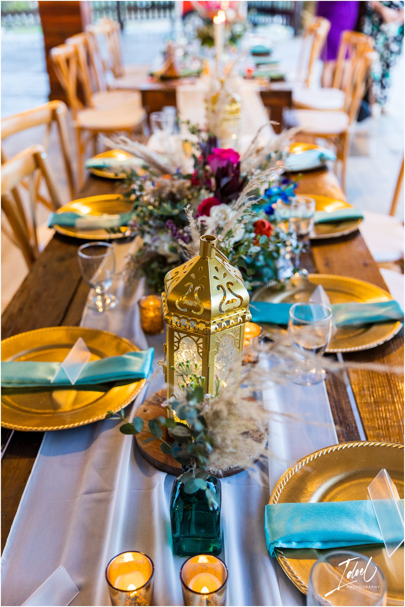 Wedding table decor featuring gold lanterns, colorful flowers, and teal accents on a rustic wooden table.