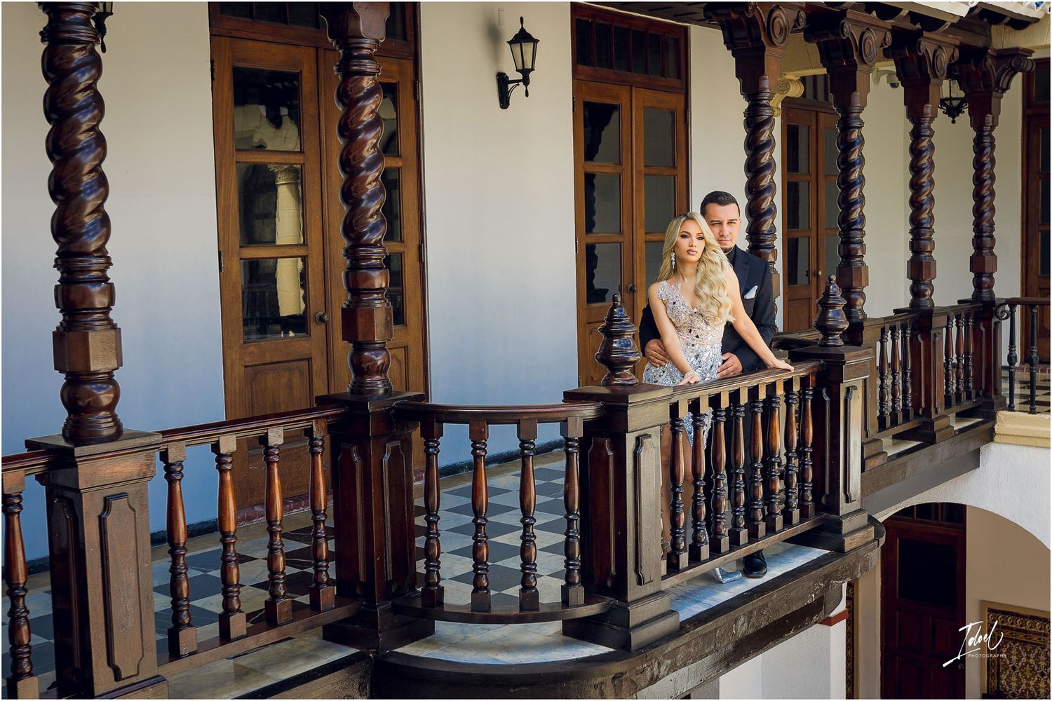 Couple poses on ornate wooden balcony of Spanish colonial building.