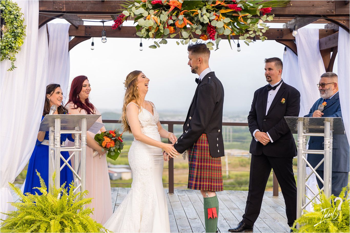 A wedding ceremony under a decorated pergola with white drapes and colorful floral arrangements.