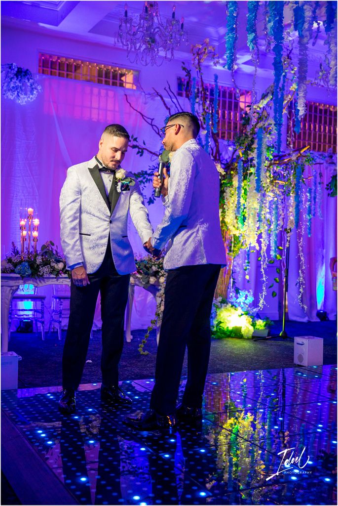 Two people in formal attire stand together at an elegant event with purple lighting and crystal chandeliers overhead.