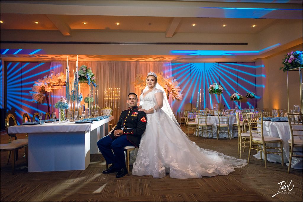 A wedding reception ballroom decorated with blue lighting and cherry blossoms features the couple at their cake table.