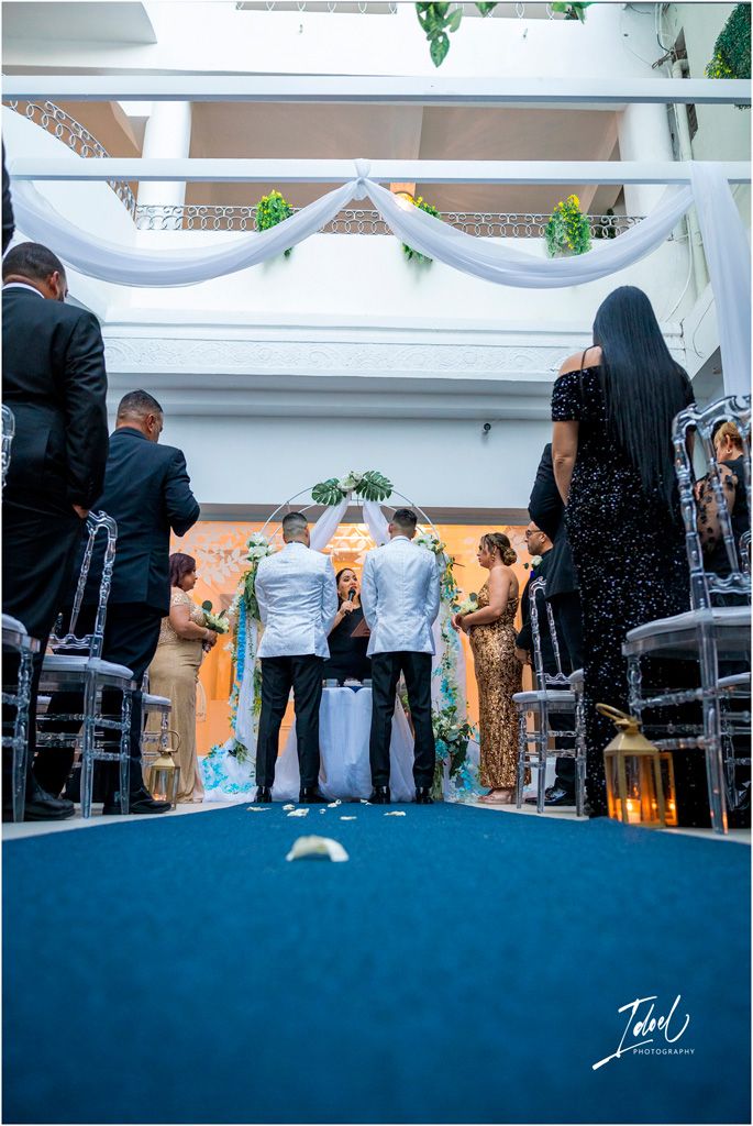 Wedding ceremony view down blue aisle runner showing couple at altar with white draped decorations and guests seated.