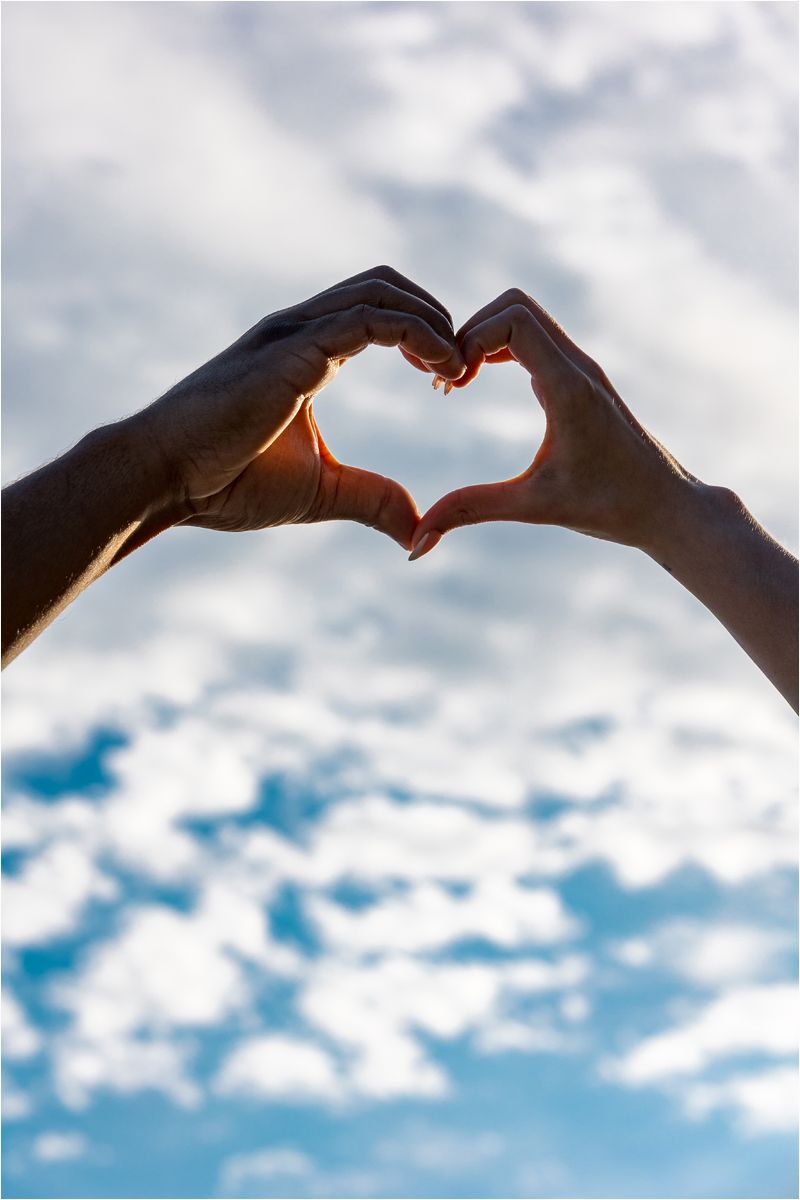 Silhouetted hands forming heart shapes against a bright blue sky with white fluffy clouds.