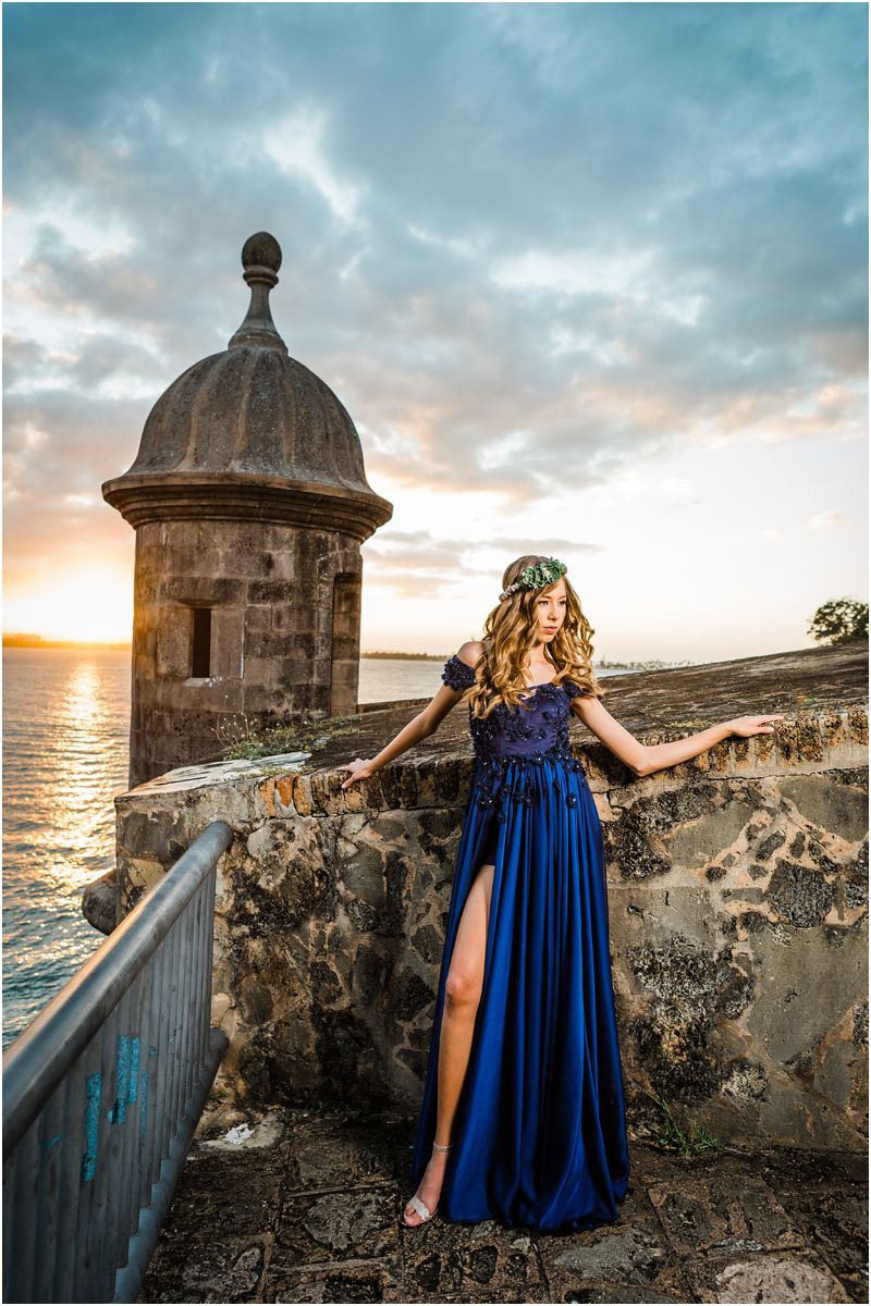 A person in a flowing blue gown poses at sunset by a historic stone watchtower overlooking the ocean.