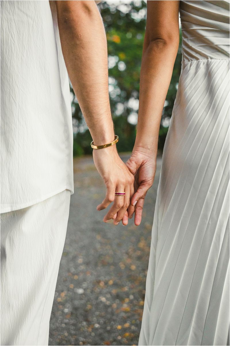 Two people in white formal attire hold hands together in an outdoor setting with a gravel path.