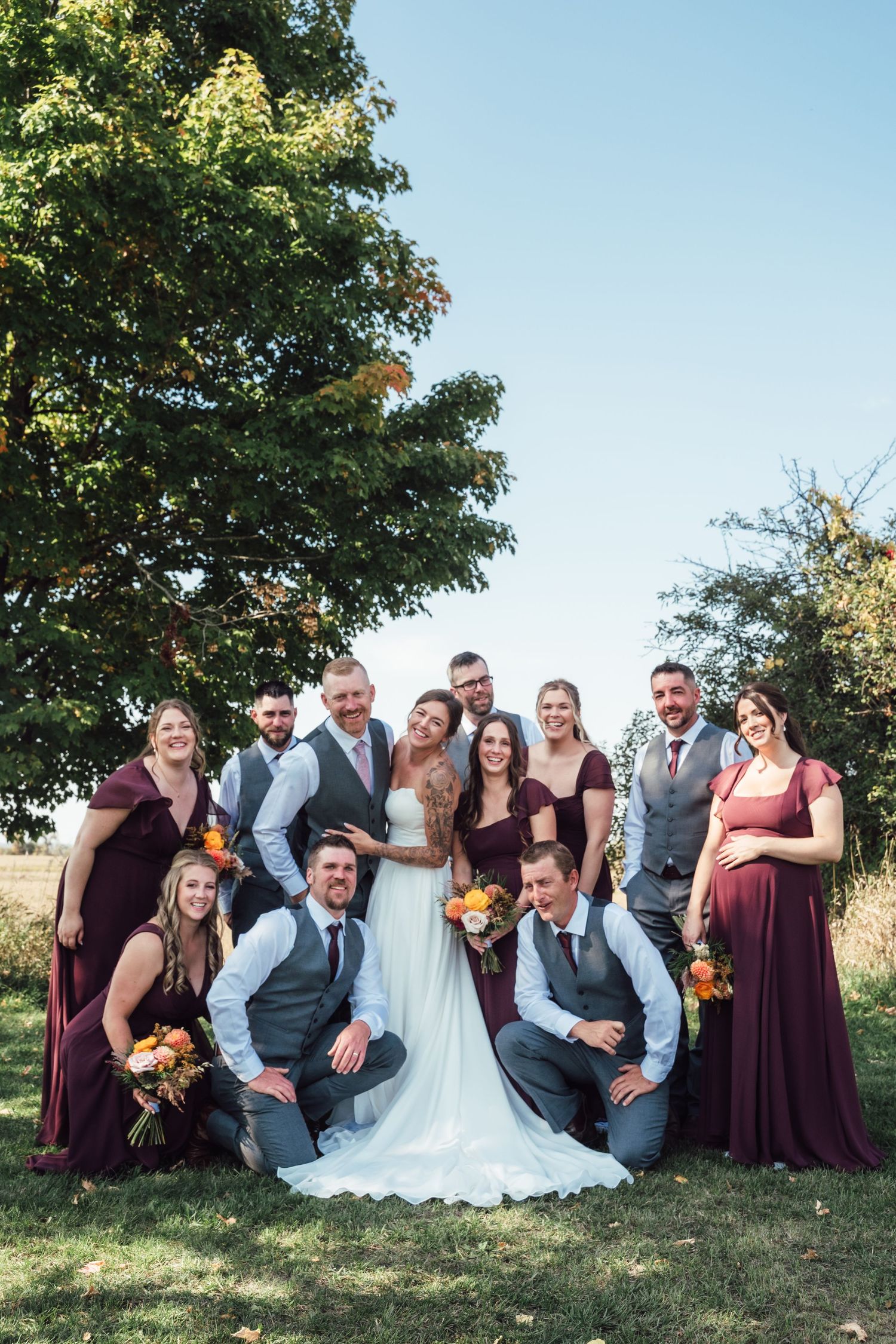 Full wedding party poses together on grass with bridesmaids in burgundy and groomsmen in gray vests.