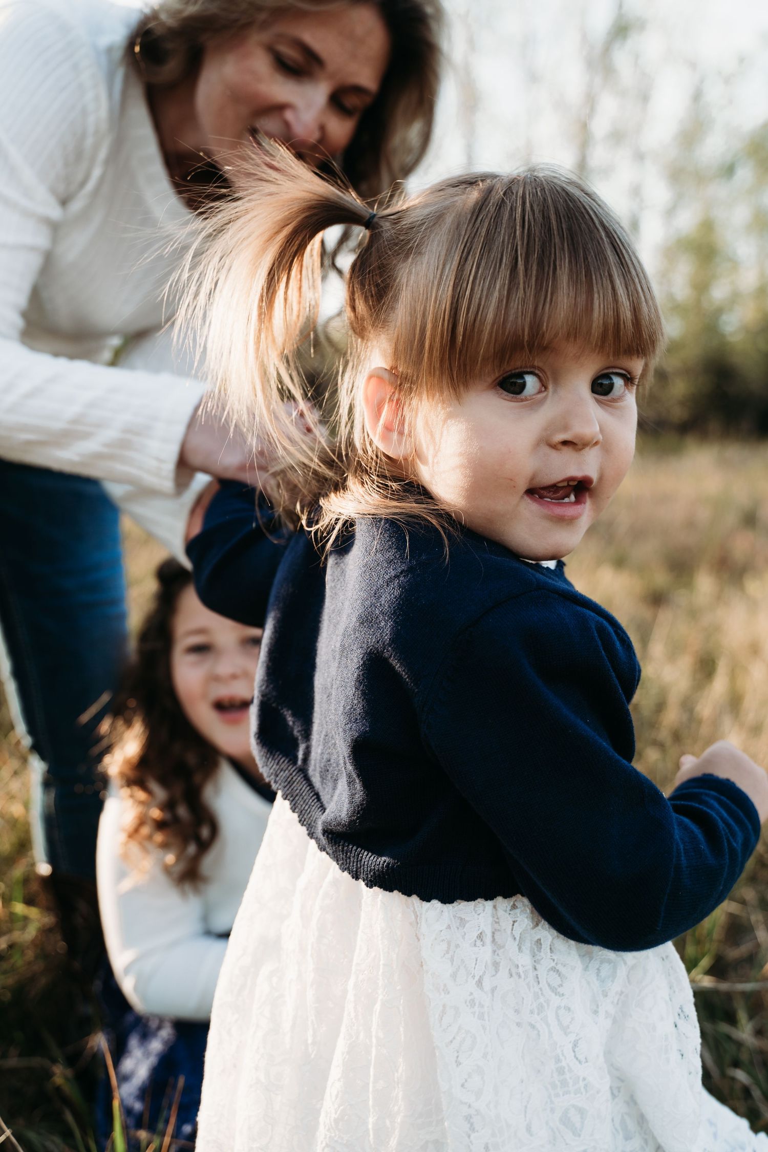 Young child in navy dress smiling playfully at the camera outdoors.