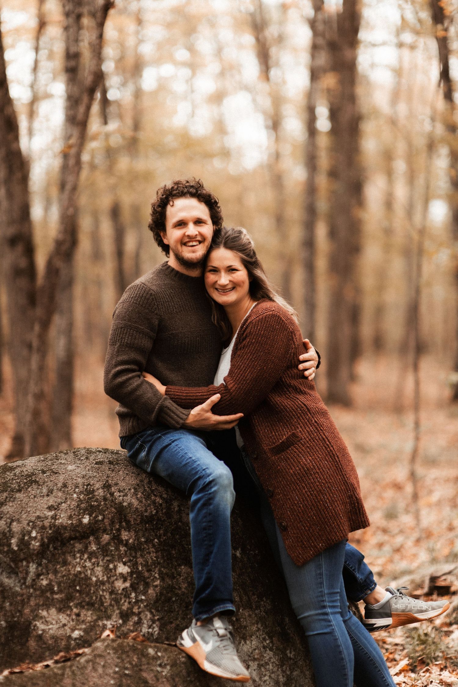 A couple shares a tender moment while sitting on a large rock in an autumn forest.