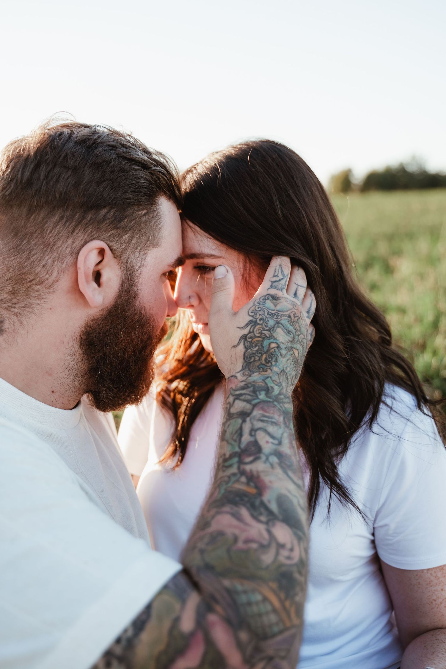 Tender moment between couple with tattooed arm in white shirts against sunset.