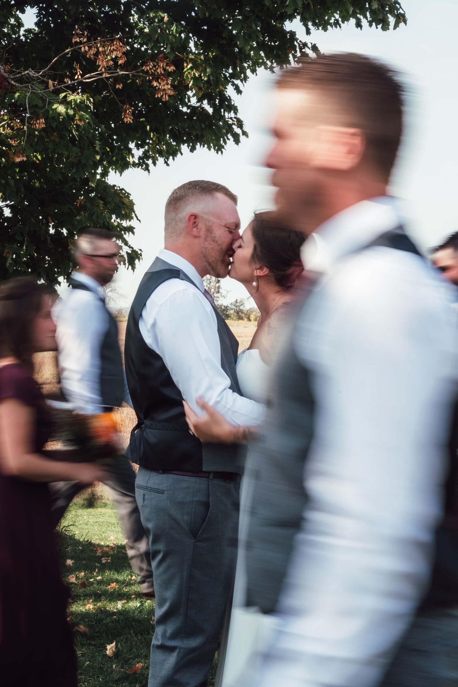 Motion blur captures guests mingling at an outdoor wedding reception under trees on a sunny day.