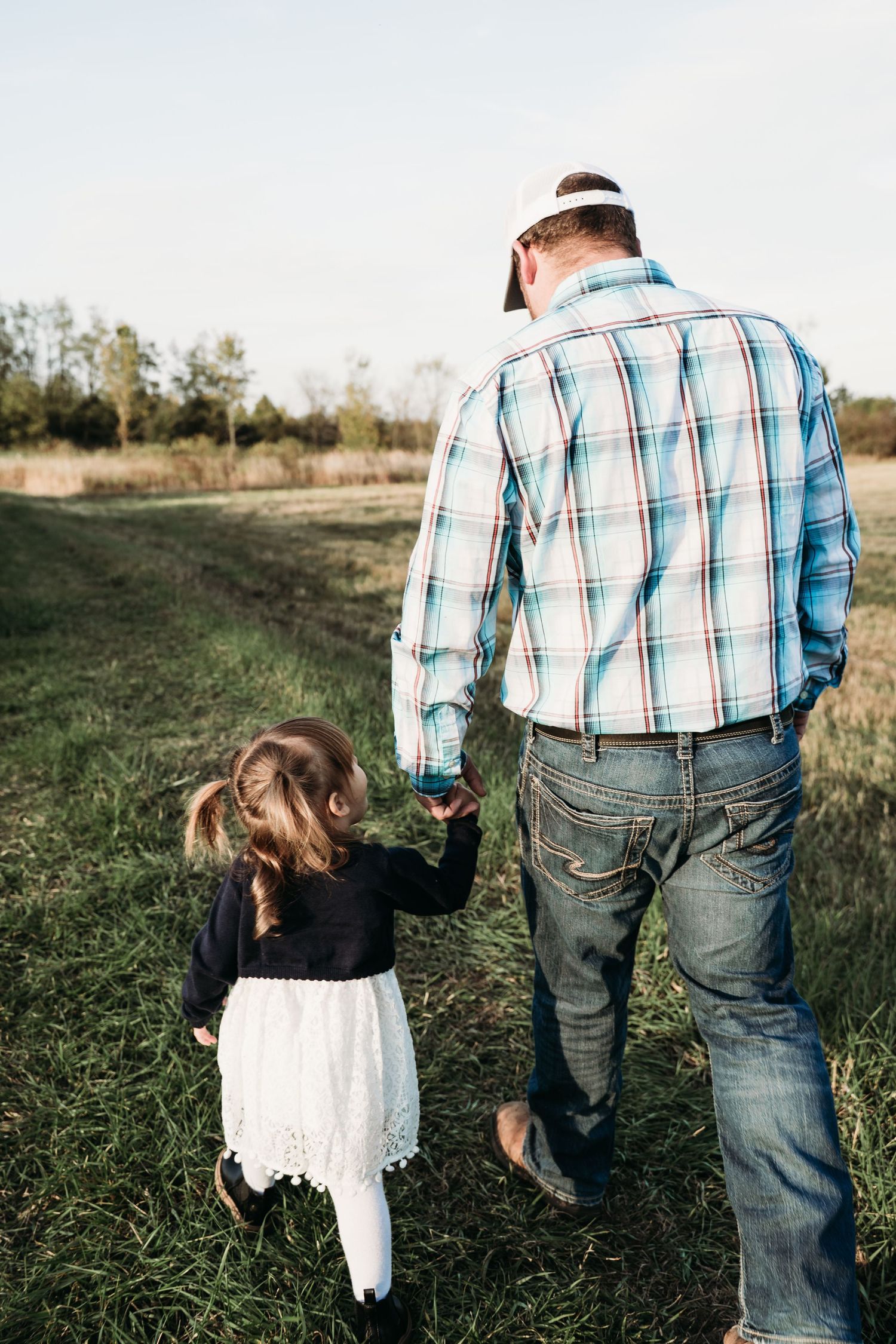 A person in a plaid shirt holds hands with a small child in a white lace dress while walking through a grassy field.