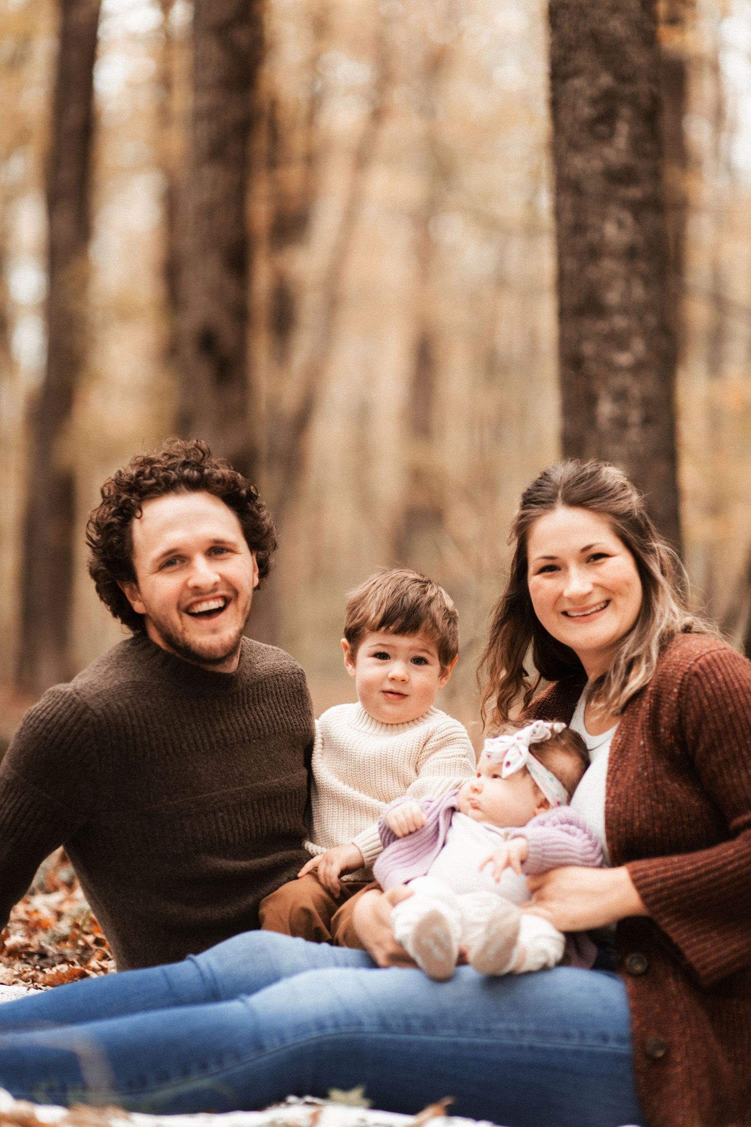 A family sits together on fallen leaves in an autumn forest for a cozy portrait.
