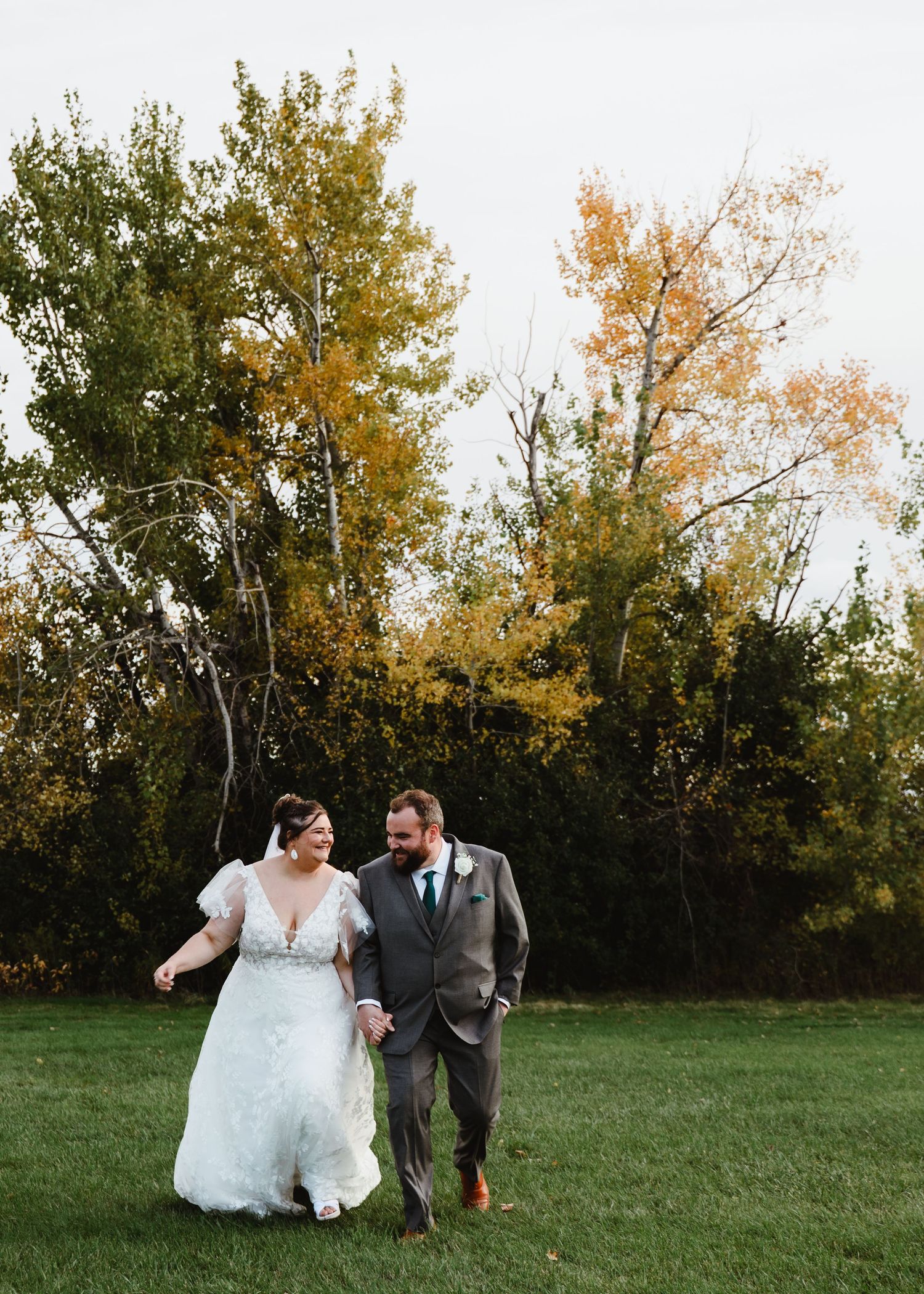 Newlyweds share playful moment while walking on grass with autumn trees in background.