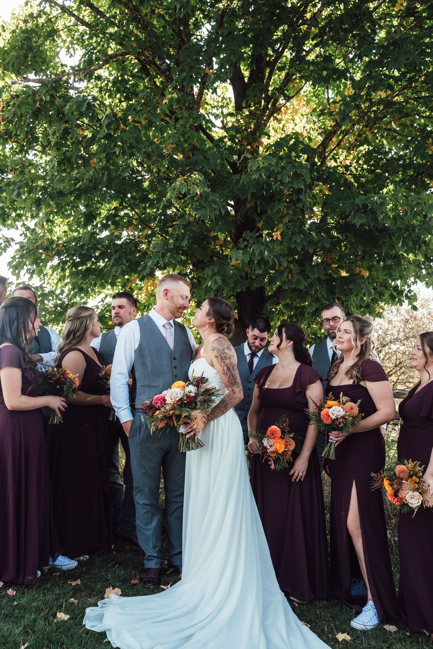 Wedding party gathers under a tree for formal photos with burgundy and white attire.