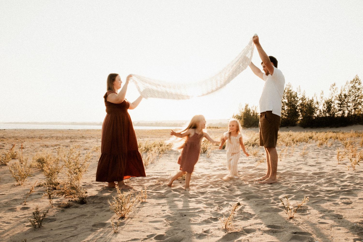 A group plays together on a sandy beach at sunset, holding a white fabric in the breeze.