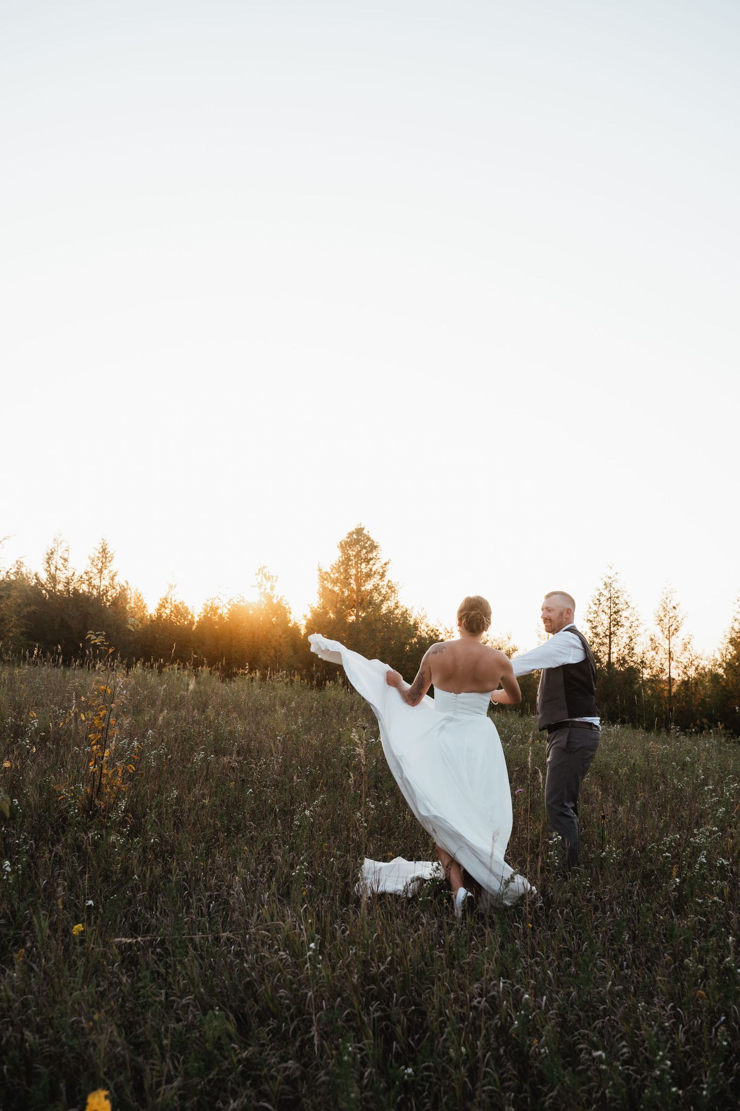 Couple poses in golden sunset light amidst tall grass with flowing white wedding dress.