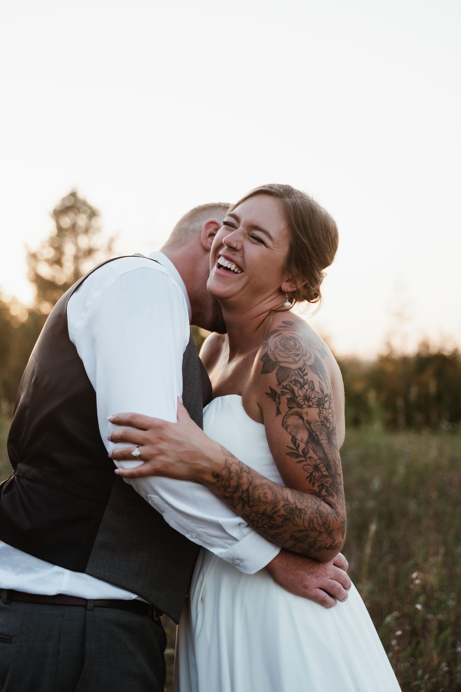 A joyful wedding moment captured in golden hour light with a tattooed bride in white dress.