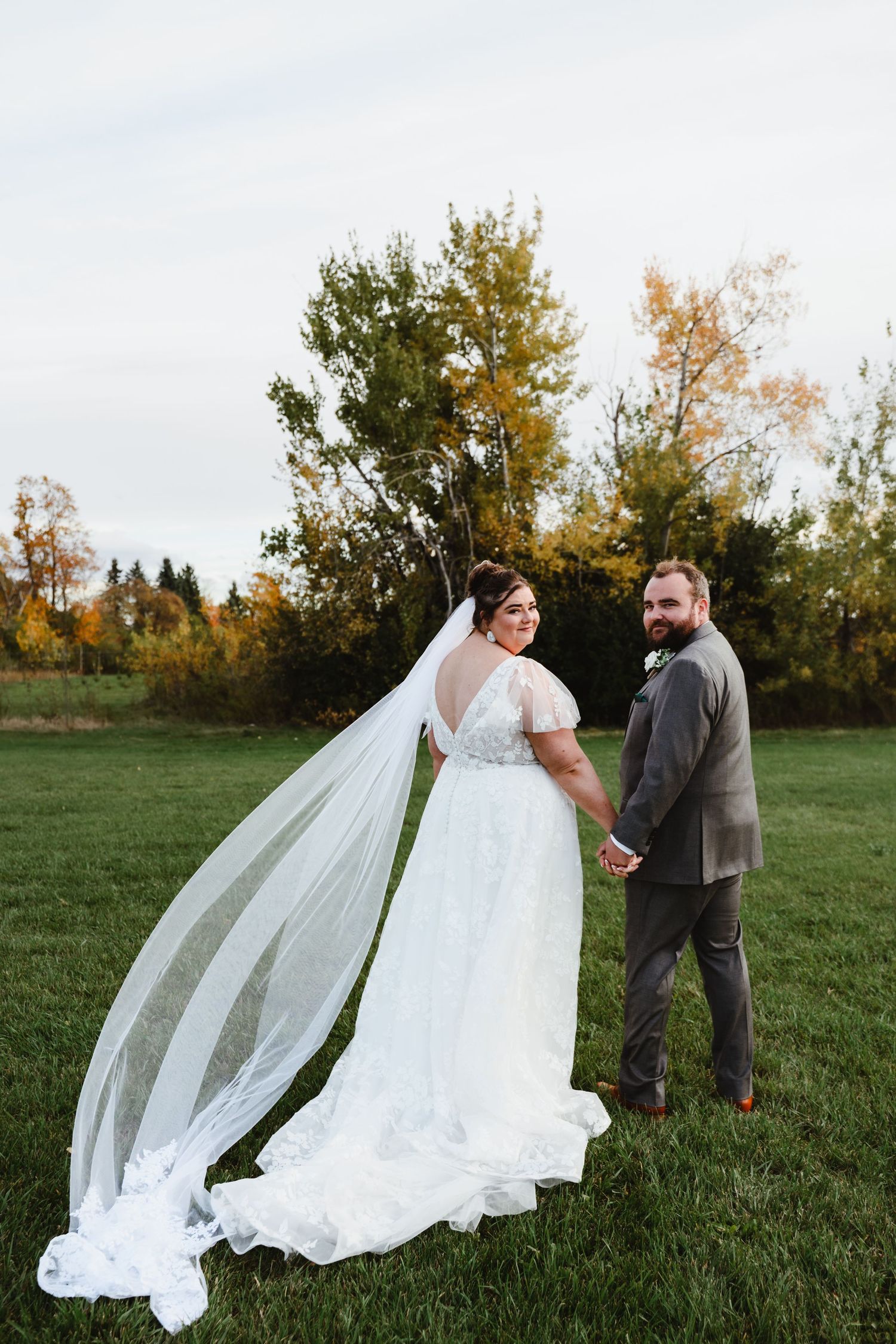 Wedding couple walks through grassy field as bride's veil flows in the wind.