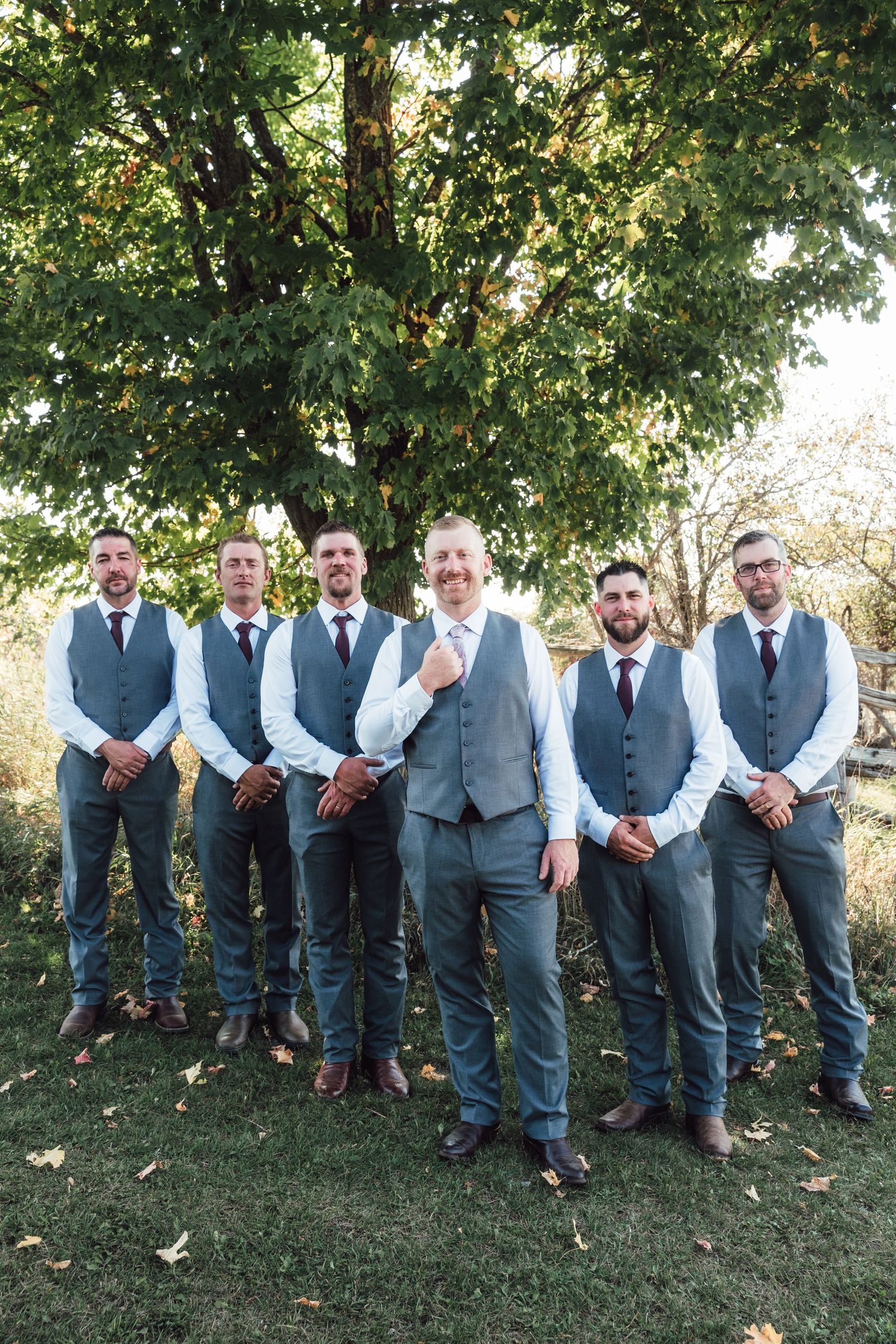 Groomsmen in matching gray vests and suits pose together under a leafy green tree.