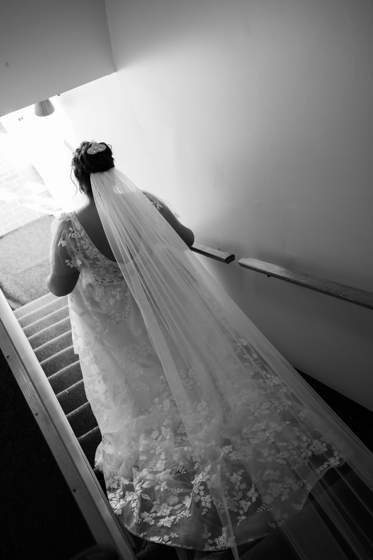 Black and white photo of bride descending stairs in detailed lace wedding gown.