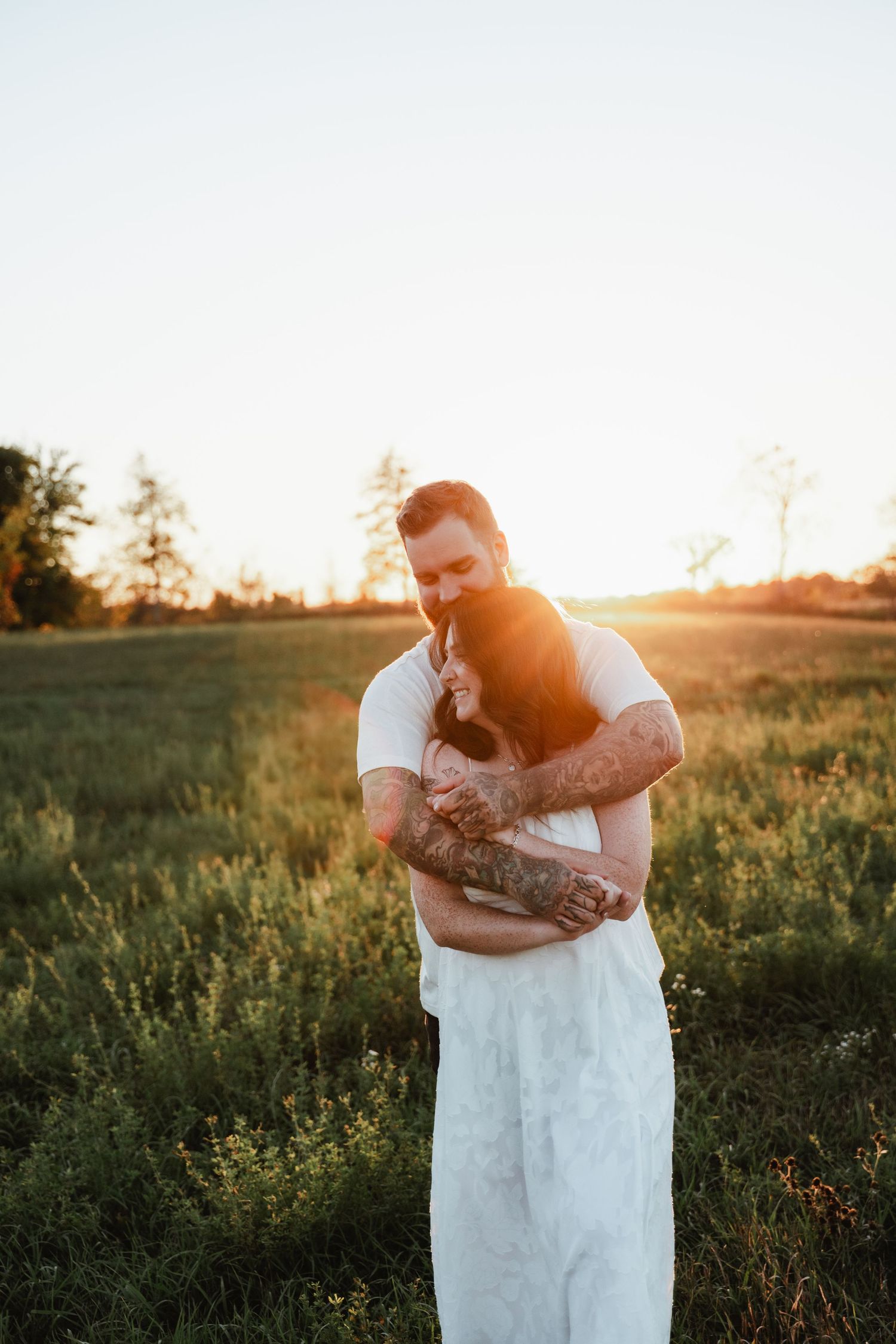 Tender embrace between couple in white clothing during sunset in grassy field.