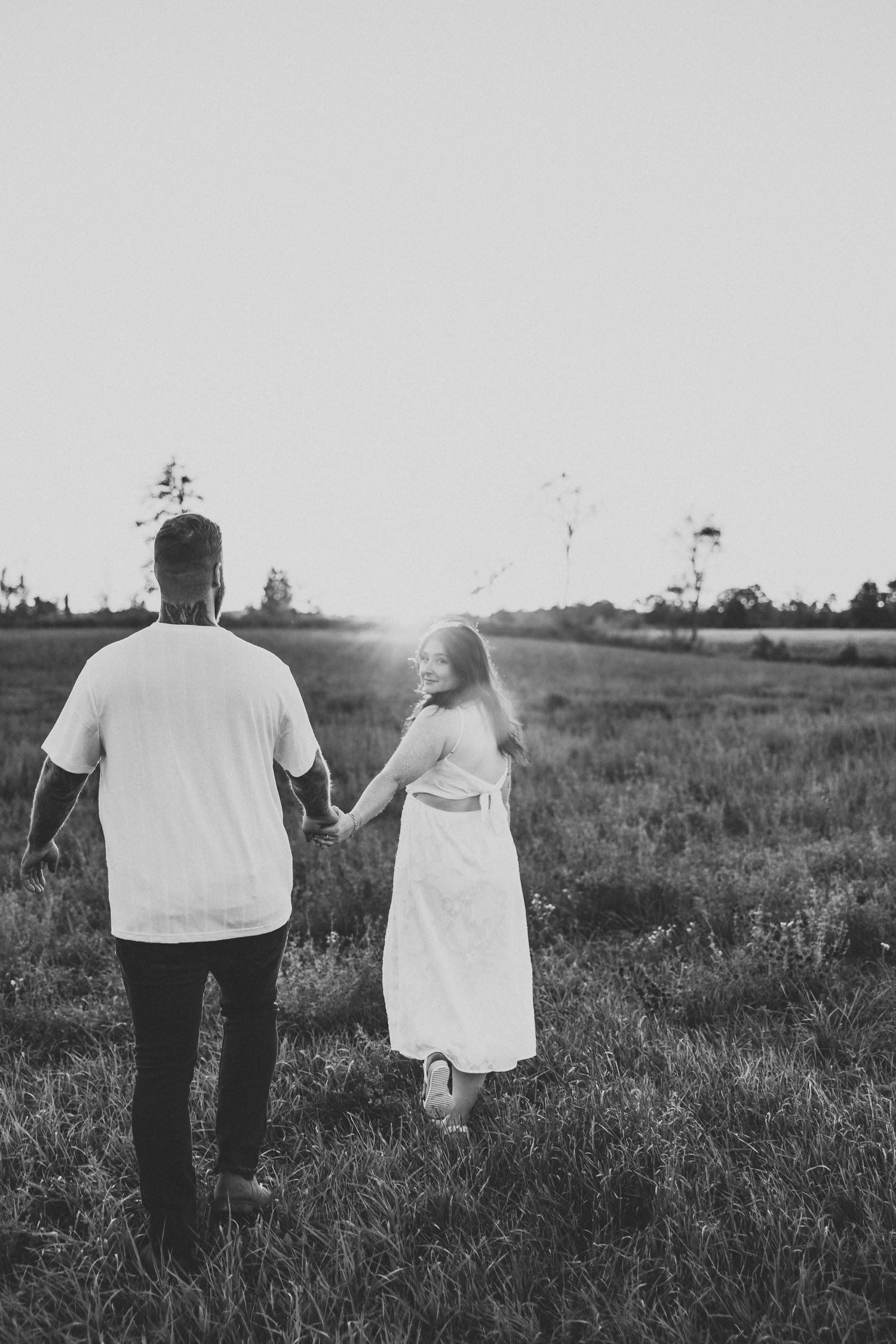 Black and white photo of couple holding hands while walking through field at sunset.
