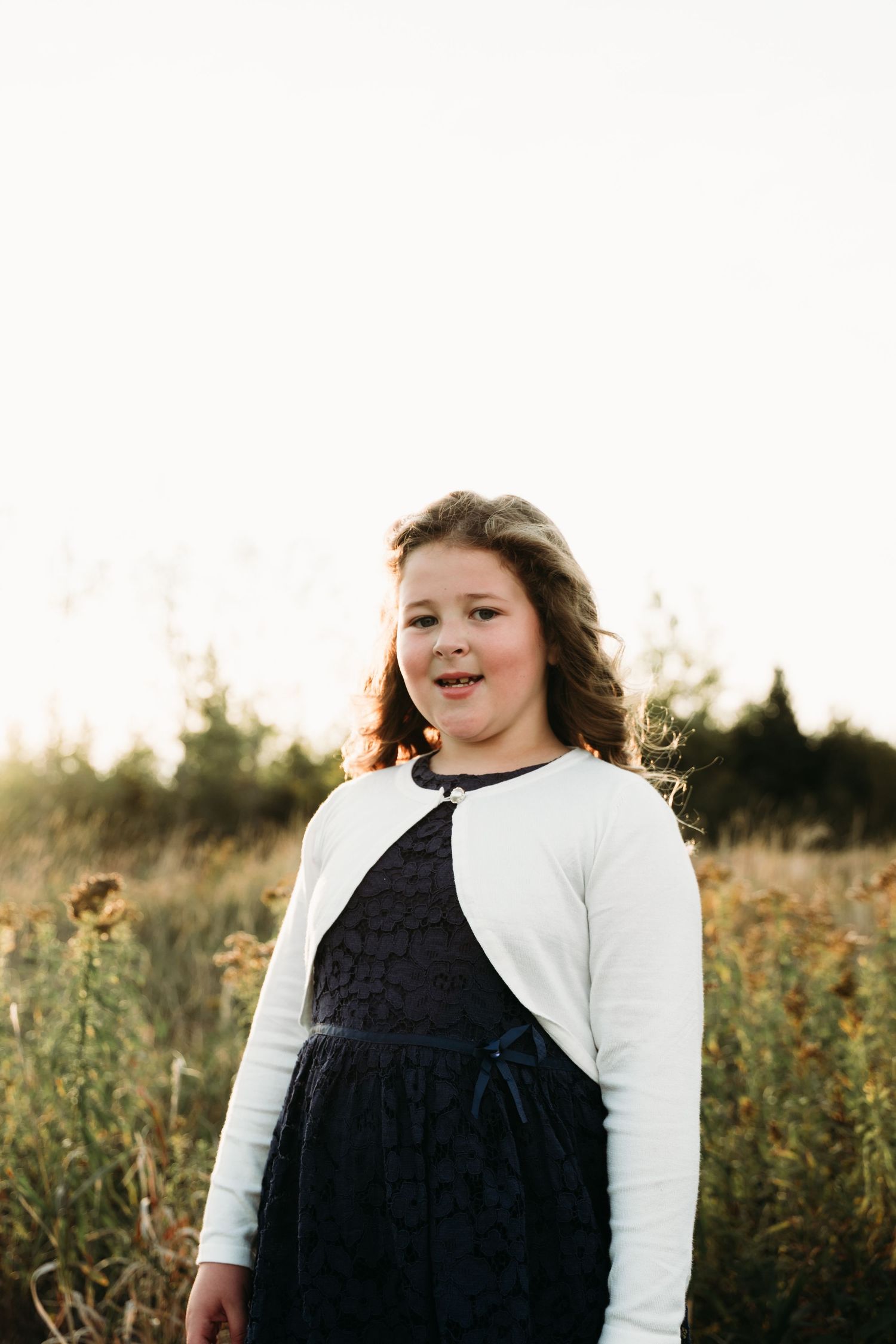 A young person in a white cardigan and navy dress stands in a sunlit field of wildflowers at golden hour.
