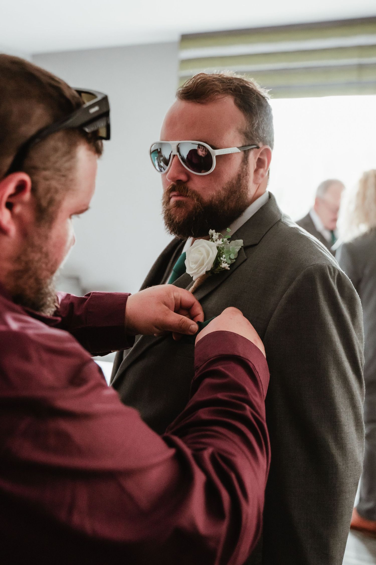Wedding party members in burgundy and grey attire adjusting boutonnieres.
