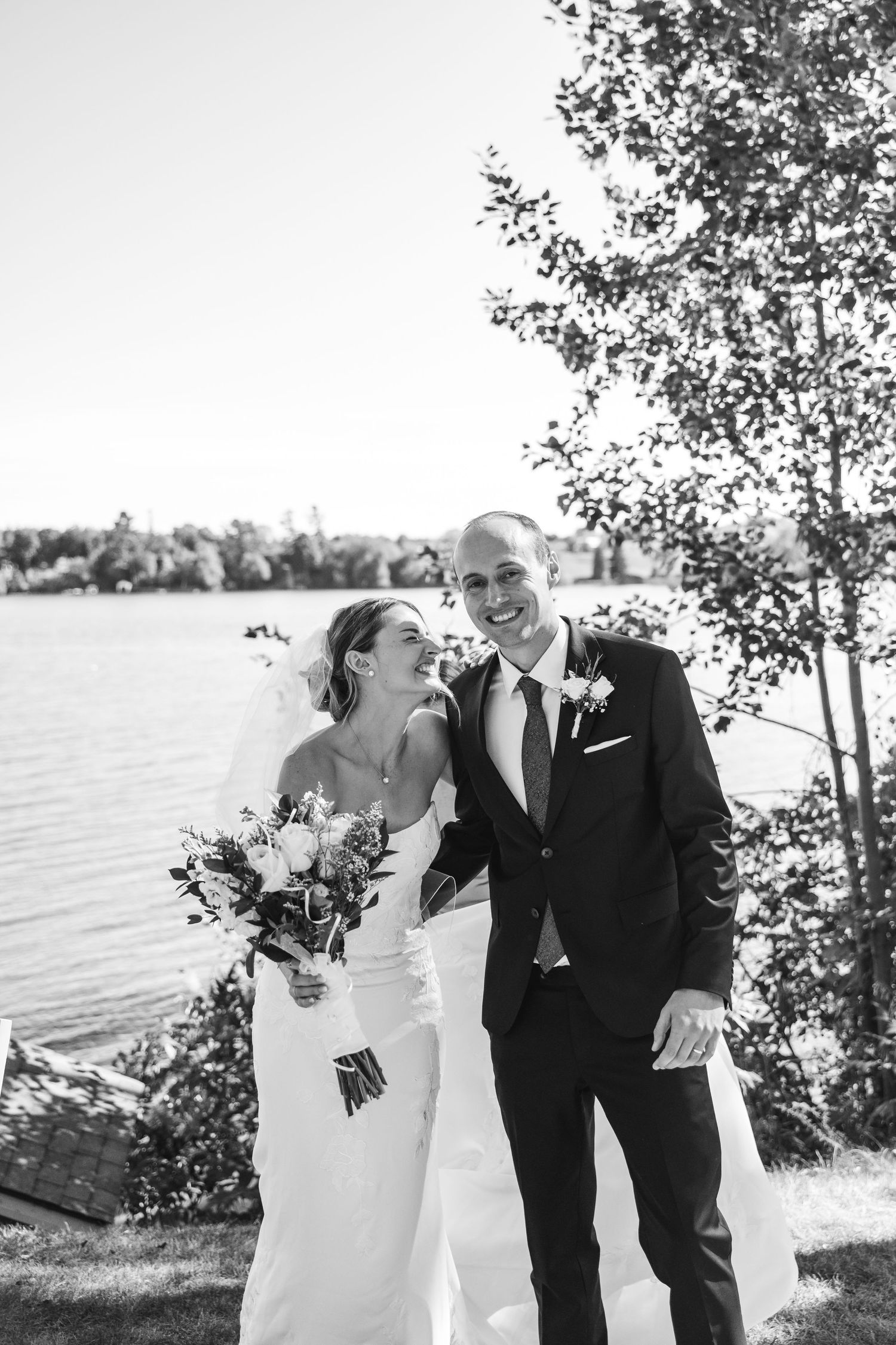 A joyful couple shares candid laughing moments by a lakeside on their wedding day.