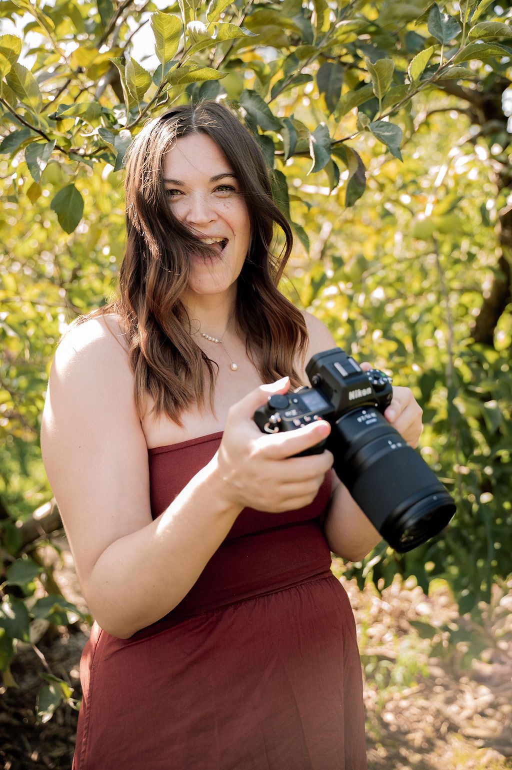 A photographer in a burgundy top checks images on their DSLR camera while standing in front of autumn foliage.