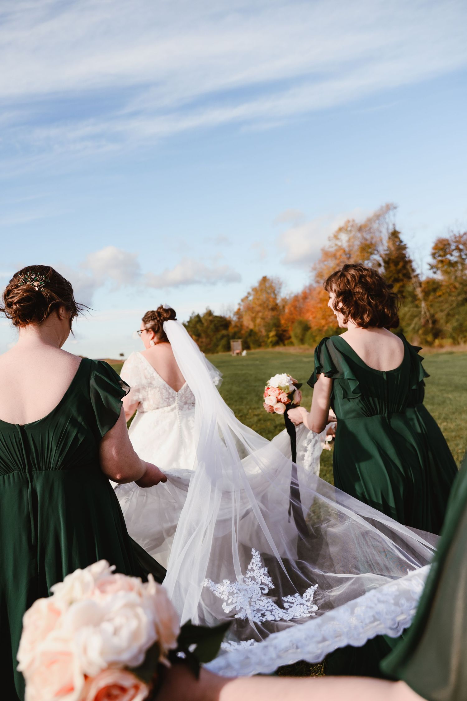 Bridesmaids in dark green dresses helping arrange the bride's lace train outdoors.