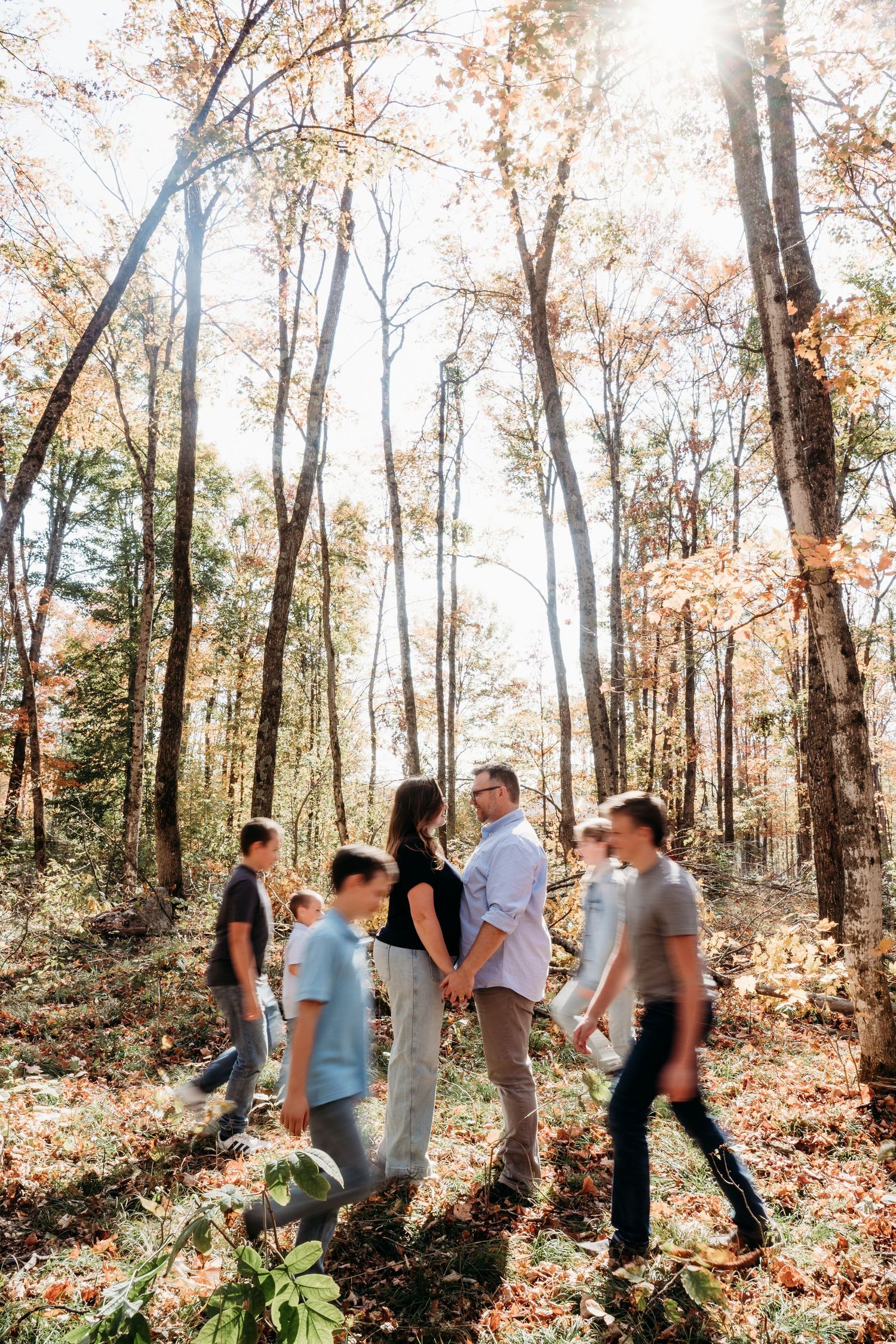 A family walks together through a sun-dappled autumn forest with fallen leaves.