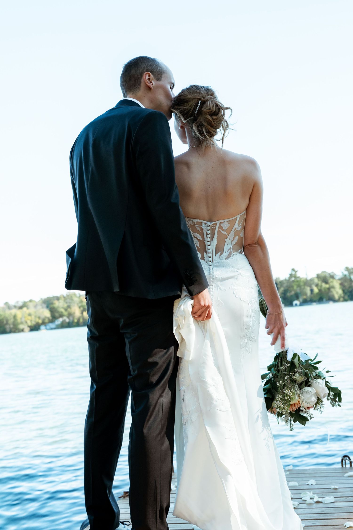 Newlyweds standing on dock overlooking blue water in elegant wedding attire.