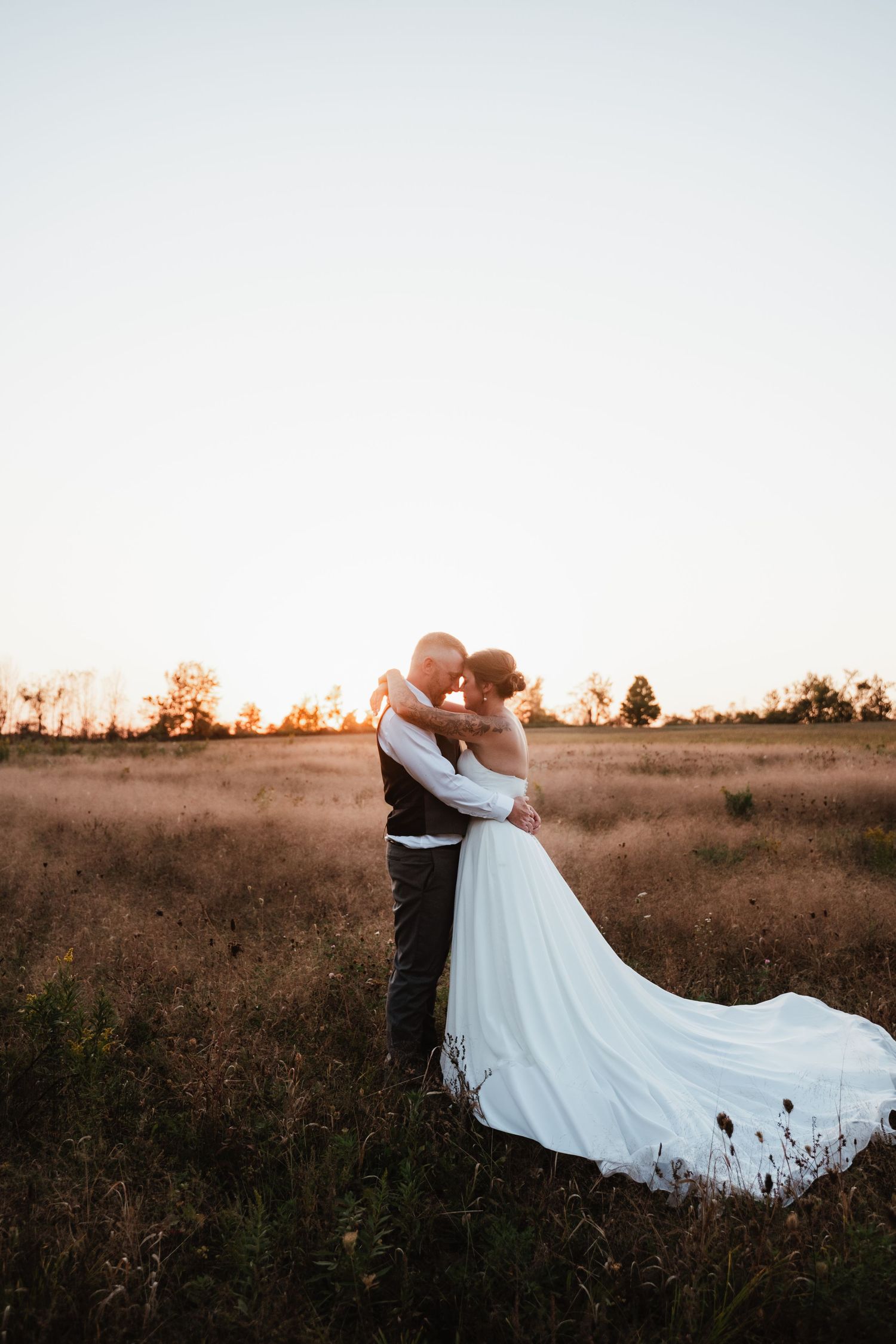 Newlyweds embrace in a rustic field during golden hour with a flowing white wedding dress.