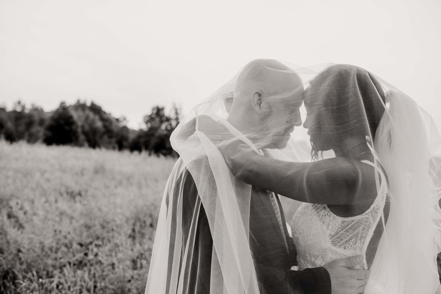 A tender black and white wedding moment captured outdoors in a field with flowing veils and natural lighting.