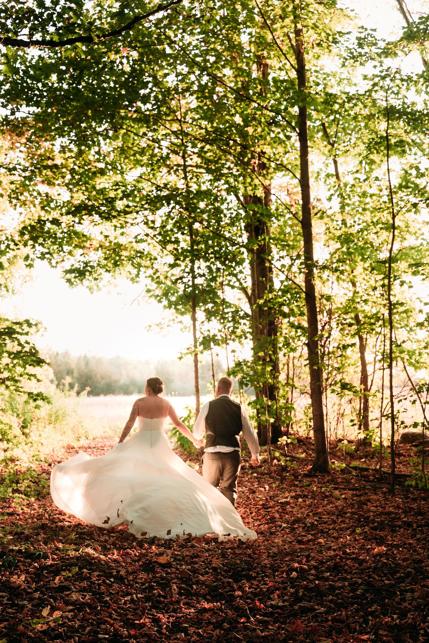 Couple walks through autumn woods as sunlight filters through trees onto wedding dress.