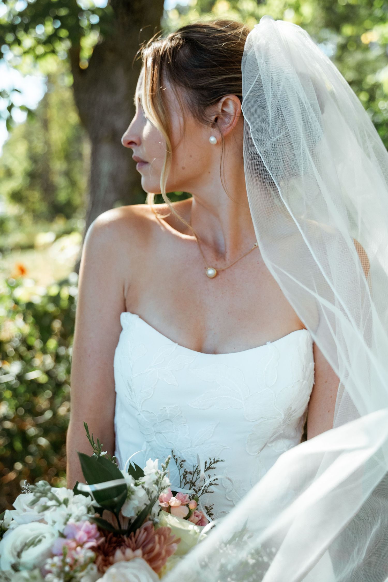 Elegant bride in strapless gown with veil holding pastel bouquet in garden setting.