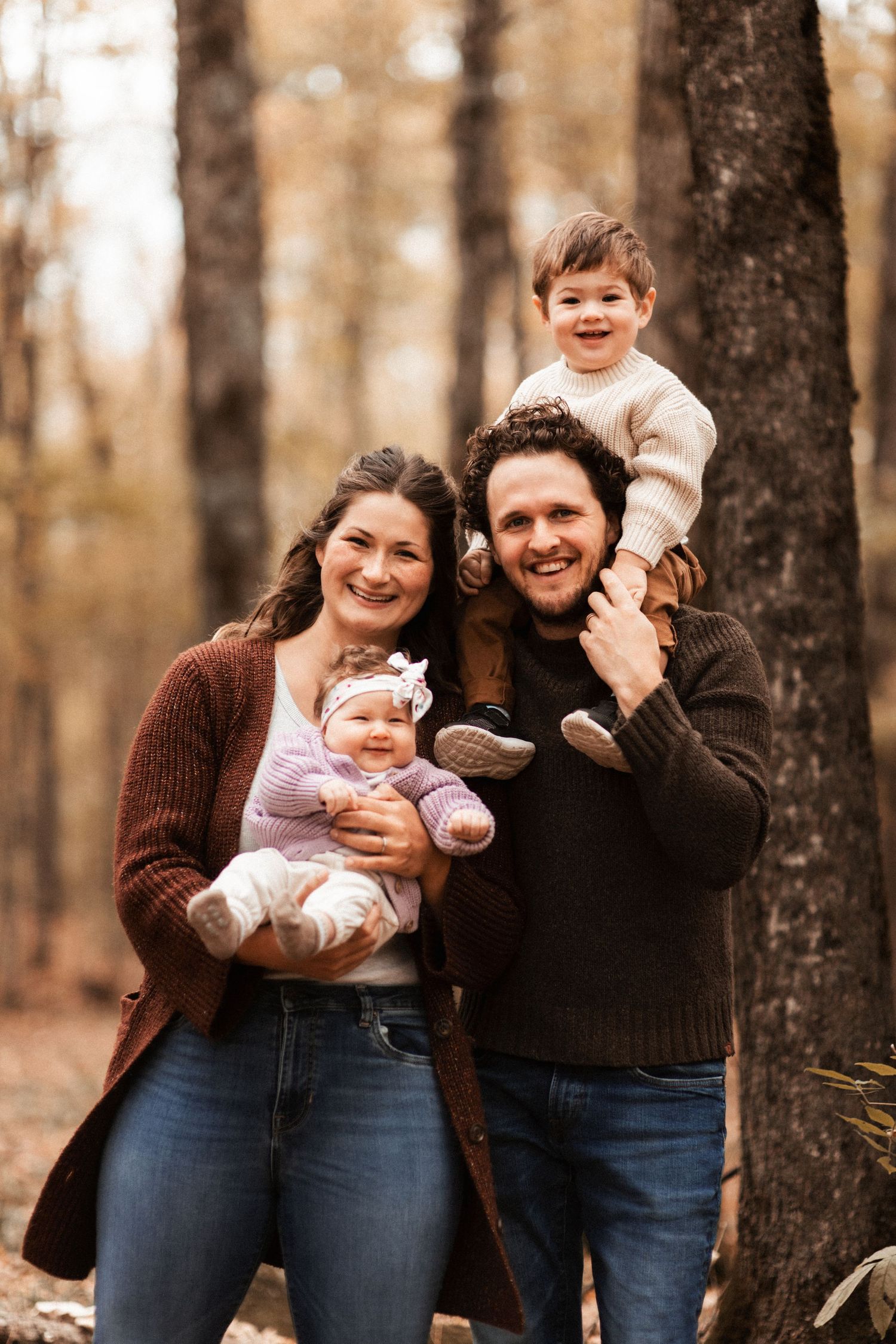A family shares warm smiles while posing together in an autumn forest setting.